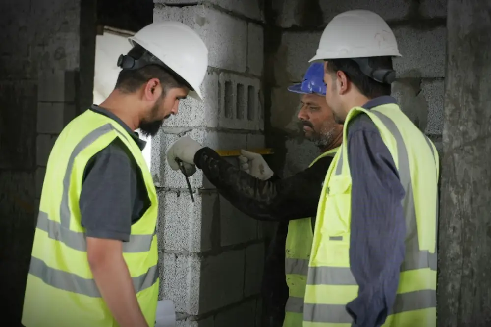 Three construction workers in high-visibility vests and hard hats measuring a concrete block wall, illustrating construction